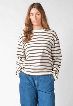 Žena s dlhými vlnitými vlasmi, oblečená v bielej a hnedo pruhovanej košeli s dlhým rukávom a modrých džínsach, ruky v vreckách, na jednoduchom pozadí.