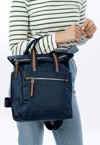 Tumesinine nailon seljakott pruunide nahkaelementidega, millel on kaks esimest lukku, kaks ülemist käepidet ja õlapaelad, struktureeritud disain.