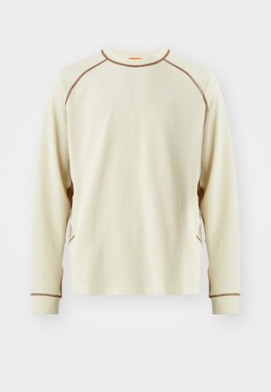 Cream long-sleeve shirt with a textured, waffle pattern. Features brown accent stitching and a small white logo on the chest.