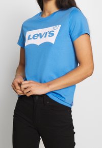 Blaues Baumwoll-T-Shirt mit einem großen weißen Levi's-Logo in der Mitte. Mit kurzen Ärmeln und Rundhalsausschnitt. Kombiniert mit schwarzen Jeans.