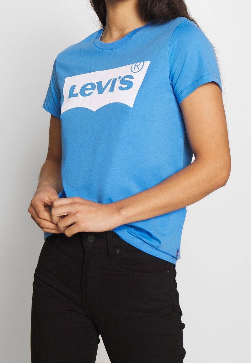 Blaues Baumwoll-T-Shirt mit einem großen weißen Levi's-Logo in der Mitte. Mit kurzen Ärmeln und Rundhalsausschnitt. Kombiniert mit schwarzen Jeans.