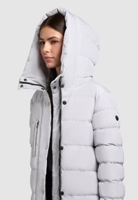 Gaiši pelēka pūdera jaka ar kapuci, ar šūtiem laukumiem, priekšējo rāvējslēdzi, spraudņiem un sānu kabatu priekšpusē.