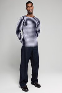 Marineblau- und weiße gestreifte Langarmbluse mit weitem Ausschnitt, kombiniert mit lockeren dunklen Jeans und schwarzen Schuhen, vor einem schlichten Hintergrund stehend.