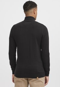 Černý žebrovaný rolák s dlouhými rukávy, s přiléhavým střihem a malou hnědou cedulkou na lemu, vyrobený z hladké pletené látky.