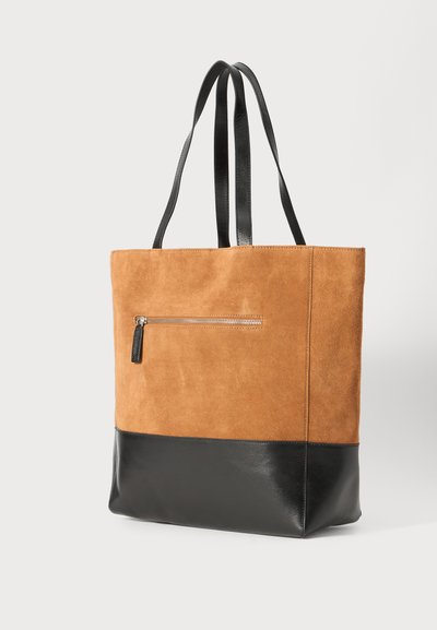 Geantă tote din suede bej cu un fund din piele neagră și un buzunar cu fermoar. Prezintă două mânere negre și o textură netedă pe suprafețe.
