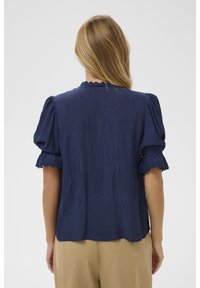 Ženska z dolgimi blond lasmi, oblečena v mornarsko modro teksturirano bluzo s kratkimi napihnjenimi rokavi in čipkastim robom, v kombinaciji z bež hlačami, obrnjena stran.