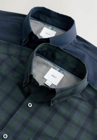 Due camicie a bottoni sovrapposte; una di colore blu navy solido e l'altra a quadri verde scuro. Entrambe presentano dettagli sul colletto e l'etichetta "NEXT" all'interno.