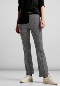 Person wearing black shirt, black-and-white patterned pants, and white sneakers standing against a plain gray background.