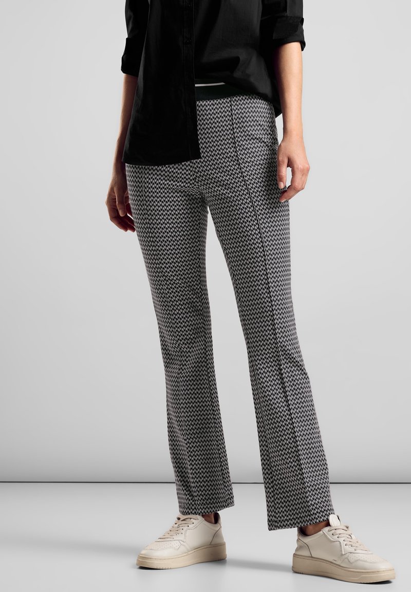 Person wearing black shirt, black-and-white patterned pants, and white sneakers standing against a plain gray background.