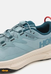 Svetlo modri trail čevlji z strukturiranim mrežastim zgornjim delom, GORE-TEX oznako, odsevnim sistemom vezalk in Vibram gumijastim podplatom.