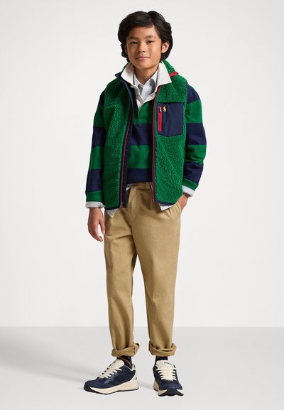 Πράσινο και ναυτικό ριγέ πουκάμισο μακρύ μανίκι, σε συνδυασμό με ένα πράσινο φλις γιλέκο με τσέπη και κόκκινο φερμουάρ, συνδυασμένο με καφέ παντελόνι και ναυτικά αθλητικά παπούτσια.