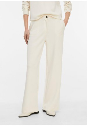 Osoba nosząca kremowe spodnie o wysokim stanie i szerokich nogawkach z bocznymi kieszeniami i zapięciem na guzik, w połączeniu z dopasowanymi kremowymi butami.