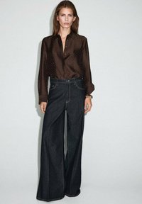 Blusa marrón con lunares y cuello, combinada con unos vaqueros oscuros de tiro alto y pernera wide-leg. Presenta un diseño ajustado y costuras visibles.