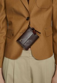 Hnědá kožená taška se stahovací šňůrkou, upevněná v pase. Taška má hladký povrch a je připevněná k světle hnědé bundě.