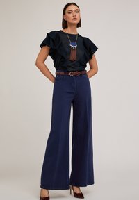 Marineblauwe wijde broek met een gladde textuur, aangevuld met een donkere ruffle-top en een bruine riem. Bevat een hangerketting met franjes.