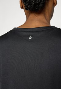 Melna sporta krekls ar teksturētu audumu un apaļu kakla izgriezumu, ar mazu logotipu aizmugurē pie apkakles.