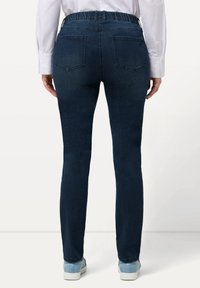 Tmavě modré džíny z denimu se strečovým pasem, slim fit a dvěma zadními kapsami. Nosič má na sobě světle modré tenisky.