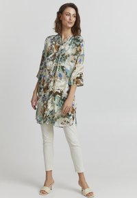 Blommig tunika i beige, gröna och blå nyanser, med trekvartsärmar och krage, tillsammans med vita slim-fit byxor och ljusa skor.