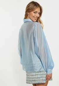 Blusa azzurra trasparente con maniche lunghe a sbuffo e colletto, abbinata a una mini gonna a motivi multicolori, che mostra una texture a strati.