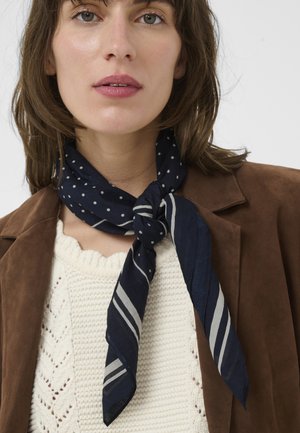 Frau trägt eine braune Jacke, einen weißen Strickpullover und einen marineblauen Schal mit weißen Punkten und Streifen, der um ihren Hals gebunden ist.