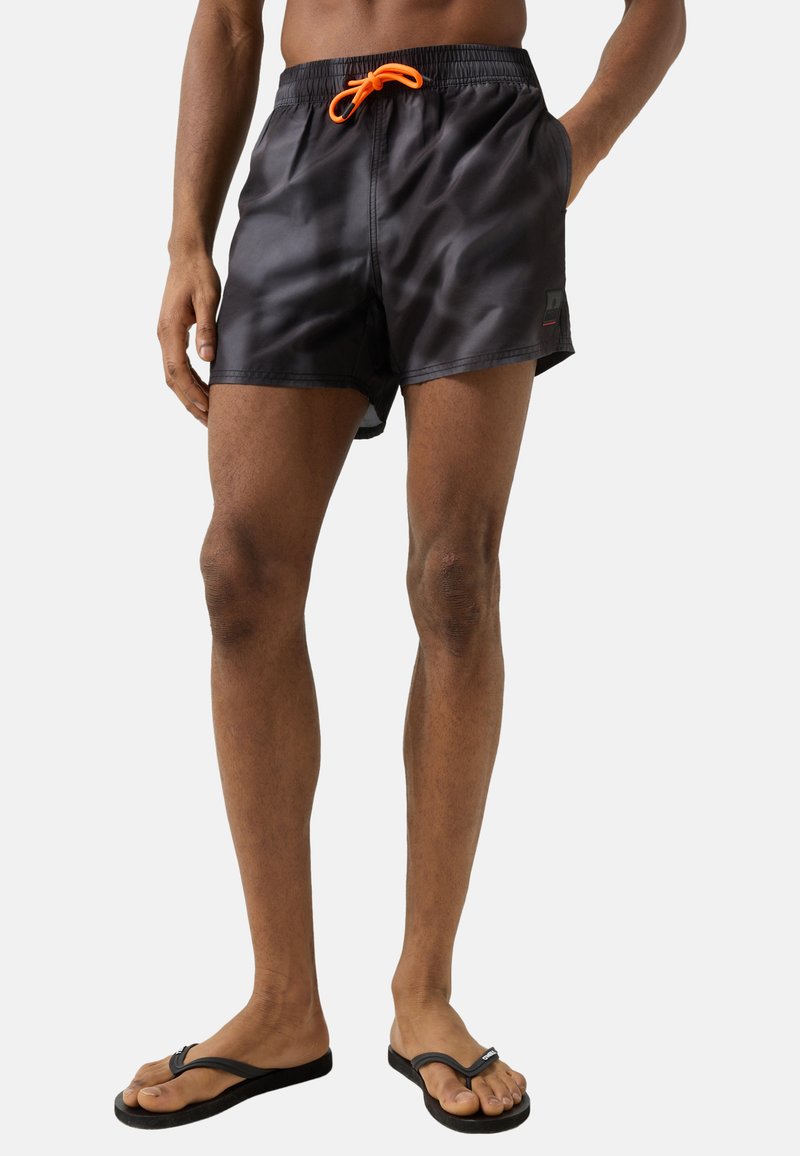Homme portant un short de bain noir avec un cordon orange et des tongs noires, debout devant un fond uni.