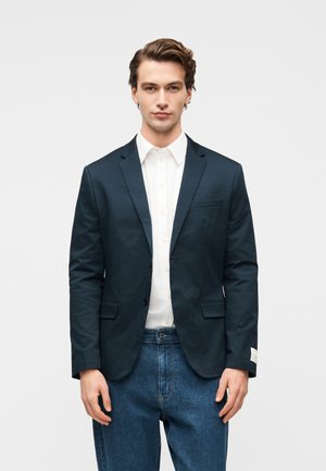 Marineblå blazer i glatt stoff, med en knapp, hakkede slag og to frontlommer. Brukes over en hvit skjorte og blå jeans.