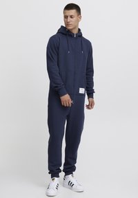 Mono con capucha azul marino con cremallera completa, bolsillos delanteros y puños de canalé. Presenta una etiqueta de marca en el bolsillo delantero.
