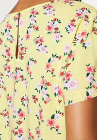 Žlutá látka s růžovým a červeným květinovým vzorem na tričku s krátkým rukávem, ukazující zadní výstřih s malým zapínáním na knoflík.