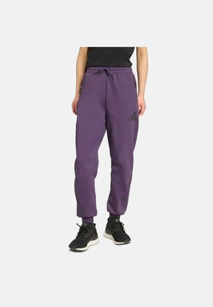 Persoon draagt paarse Adidas joggers met trekkoord in de taille en zwarte sneakers, staand tegen een effen lichte achtergrond.