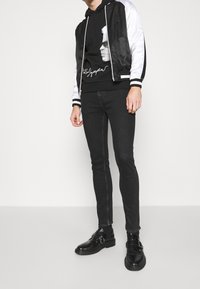 Chaqueta bomber negra con mangas plateadas sobre una sudadera negra con gráfico, combinada con pantalones ajustados negros y botas negras con hebilla. Diseño minimalista.