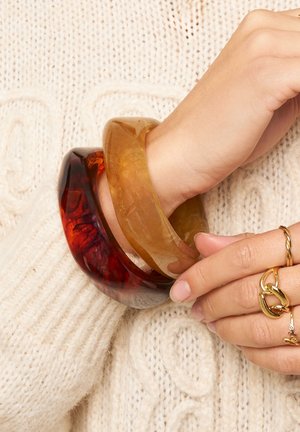 Drei Armbänder: eines in Bernsteinfarbe, eines transparent mit orangefarbener Tönung und eines dunkelrot, alle mit glatten, glänzenden Oberflächen.