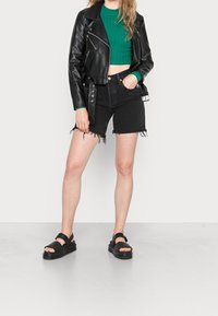 Schwarze, cropped Motorradjacke aus Leder, grüner gerippter Pullover, schwarze Jeansshorts mit fransigem Saum und schwarze Slipper mit verstellbaren Riemen.
