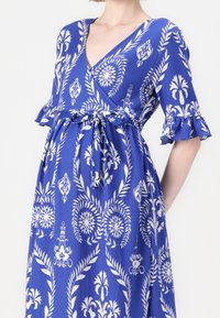 Blå omlottklänning med vita blommönster, korta volangärmar, ett bälte i midjan och V-ringning. Strukturerad tyg.