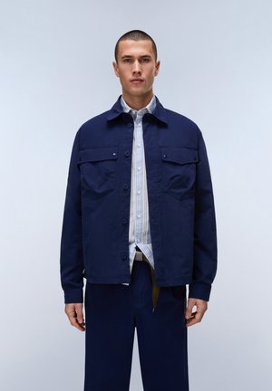 Mand iført navyblå, knappet jakke over en stribet skjorte og matchende navybukser, stående mod en ensartet lys baggrund.