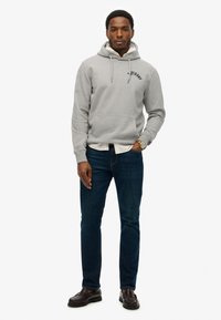 Grijze hoodie met een voorzak en trekkoorden, gedragen over een wit overhemd met kraag; gecombineerd met blauwe jeans en donkere instappers.