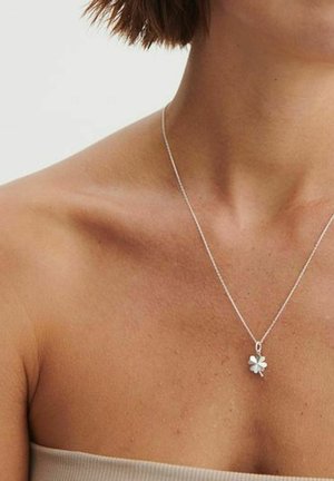 Collar de plata con un colgante de trébol de cuatro hojas, llevado en un cuello y hombros desnudos, con cabello corto y castaño visible sobre un fondo claro.