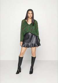 Blusa verde com padrão e decote em V, combinada com uma saia preta de couro em formato evasê. Botas pretas até ao joelho completam o look.