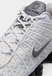 Posnetek iz bližine bele in sive superge Nike Shox, ki prikazuje teksturirano zgornji del, vezalke in črno podloženo podplatno podrobnost.