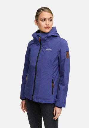 Woman wearing a blue zip-up hooded jacket with logo on chest and patch on sleeve, standing with hands relaxed by her sides.