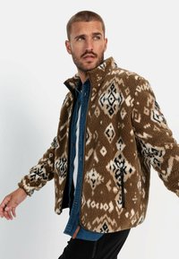 Braune Fleecejacke mit weißen und schwarzen geometrischen Mustern, mit einem Reißverschluss vorne, hohem Kragen und seitlichen Taschen. Strukturelle Oberfläche.