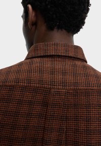 Camisa a cuadros marrón con detalles en negro. Presenta un cuello extendido y una tela texturizada, mostrando un patrón de espiga en la parte posterior.