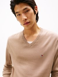 Pull en maille beige avec un col en V et des manches longues. Présente un petit logo de la marque sur la poitrine gauche. Texture lisse, coupe ajustée.