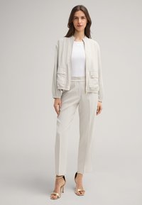 Helle beige Bomberjacke mit zwei Fronttaschen, elastischen Bündchen, einem hohen Kragen, kombiniert mit passenden tapering Hosen und goldenen Riemchen-Pumps.