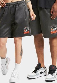 Grå fleece shorts med elastisk midje, prydet med en rød og svart "GUARDS"-logo. Kombinert med hvite joggesko og synlige tatoveringer.