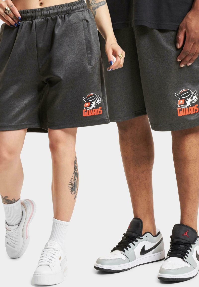 Grå fleece shorts med elastisk midje, prydet med en rød og svart "GUARDS"-logo. Kombinert med hvite joggesko og synlige tatoveringer.