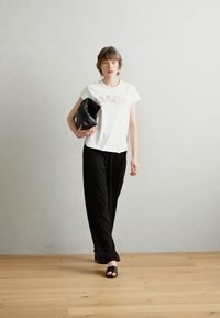 Bílé tričko s nápisem "CREATE", černé široké kalhoty a černá kabelka. Modelka má na sobě černé skluzové sandály a stojí proti světlému pozadí.