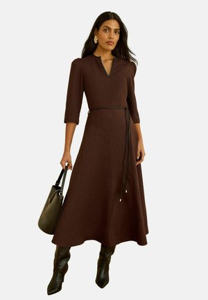 Braunes knieumspielendes Kleid mit leicht ausgestellter A-Linien-Silhouette, dreiviertellangen Ärmeln und einem Taillenzug. Kombiniert mit schwarzen kniehohen Stiefeln.