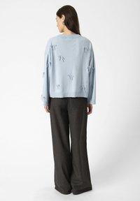 A woman stands with her back to the camera, wearing a light blue sweater adorned with bows and dark, wide-legged trousers.