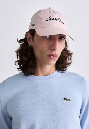 Jaunas žmogus su ilgais garbanotais plaukais dėvi rožinę Lacoste kepurę ir šviesiai mėlyną megztinį su Lacoste krokodilo logotipu ant krūtinės.