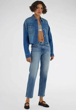 Donna che indossa una giacca corta in denim sopra un top corto grigio, jeans blu slavati a vita alta e ballerine nere Mary Jane, in piedi su sfondo bianco.